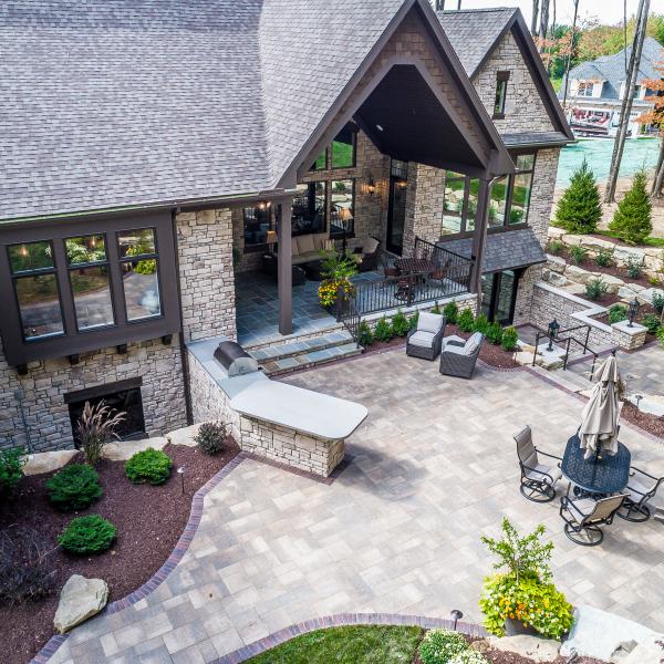 A stone patio featuring a built-in grill, multiple seating areas, and a large fireplace surrounded by expert landscaping.