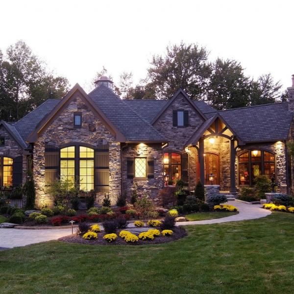 A custom-built stone cottage surrounded by beautiful landscaping lit up at night through large windows and exterior lighting.