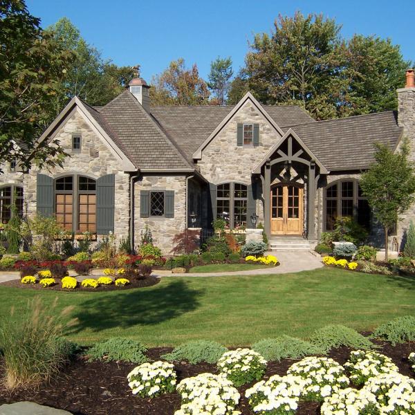 A custom-built stone cottage home surrounded by beautiful landscaping features wood shutters and large windows.
