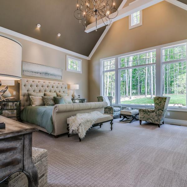 A large master bedroom featuring high ceilings and exposed beams with large windows looking out into a wooded backyard.